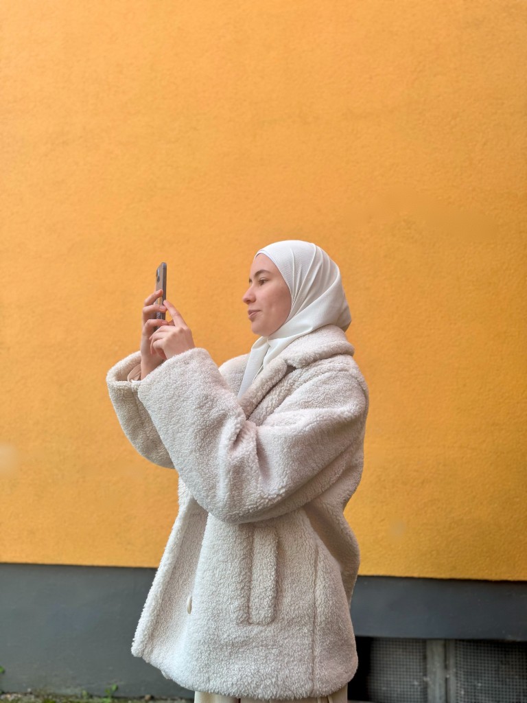 Farah aus dem delta3 Team fotografiert mit dem Smartphone vor einer gelben Wand